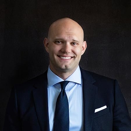 Retrato profissional de James Clear, escritor e palestrante especializado em formação de hábitos e desenvolvimento pessoal. Ele aparece sorrindo, com a cabeça raspada, vestindo um terno azul-marinho, camisa azul-clara, gravata azul-escura e lenço branco no bolso do paletó. A imagem é enquadrada da cintura para cima, com o autor olhando diretamente para a câmera e transmitindo uma expressão confiante, simpática e acolhedora. O fundo é escuro e liso, destacando sua figura e conferindo um aspecto elegante e profissional ao retrato.
