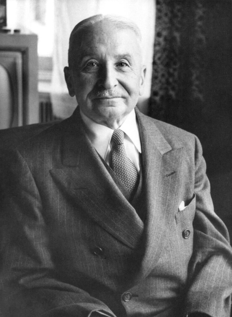 Retrato em preto e branco de Ludwig von Mises, economista austríaco e um dos principais representantes da Escola Austríaca de Economia. O autor aparece sentado, usando terno escuro e gravata, com expressão serena e olhando diretamente para a câmera. A fotografia destaca sua postura elegante e transmite a imagem de um intelectual que influenciou profundamente os estudos sobre economia, liberdade econômica e teoria monetária.