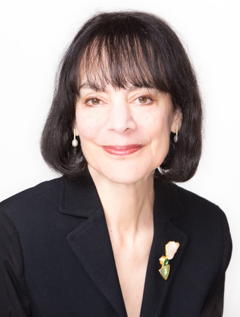 Retrato de Carol S. Dweck, psicóloga e pesquisadora norte-americana conhecida por desenvolver a teoria do mindset. Ela aparece em um enquadramento de meio corpo, olhando diretamente para a câmera com uma expressão serena e um leve sorriso. Tem cabelos escuros, lisos, na altura dos ombros, com franja sobre a testa, olhos castanhos e usa brincos discretos de pérola. Veste um blazer preto sobre uma blusa escura e um broche em formato de flor com detalhes dourados, verdes e brancos preso à lapela. O fundo é claro e desfocado, destacando a autora e transmitindo uma imagem profissional, acolhedora e elegante.